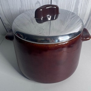May include: A round, dark brown ceramic bean pot with two side handles and a shiny, metallic silver lid. The lid features a dark brown knob handle at the centre. The pot has a smooth, glazed finish and a deep, uniform colour throughout the body.