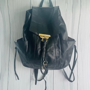 May include: Black leather backpack with a flap closure and side pockets. The backpack features gold-tone hardware, including a clasp and decorative rings. A braided tassel hangs from the front.