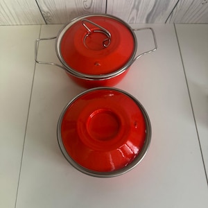 May include: Two red enamel pots with stainless steel rims and handles. One pot has its lid on, and the other has its lid off. The lids and pots are a bright, solid red color.