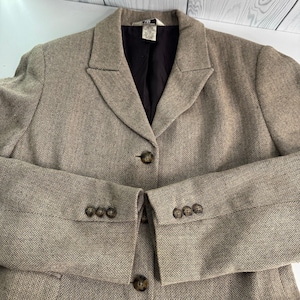 May include: A tan and brown herringbone blazer featuring a notched lapel, a dark brown interior lining, and a single-button front closure. The sleeves are finished with four small brown buttons on each cuff, and a white brand label is visible.