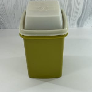 Puede incluir: Un recipiente de plástico verde oliva vintage con forma cuadrada y tapa beige claro. La tapa tiene una parte superior ligeramente empotrada con un diseño circular en el centro. El recipiente tiene una base rectangular y un borde verde oliva a juego.