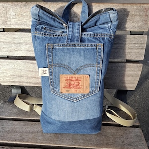 Op de afbeelding: Een blauwe denim rugzak met een rechthoekige vorm. De rugzak heeft een achterzak met een bruine leren patch met de tekst "Levi Strauss & Co." en een klein wit label. Hij heeft een handvat aan de bovenkant en verstelbare beige bandjes.
