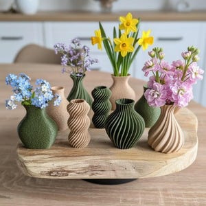 Könnte beinhalten: Eine Sammlung kleiner, dekorativer Vasen in verschiedenen Farben und Designs, die frische Blumen enthalten. Die Vasen sind auf einem Holzservierbrett angeordnet und präsentieren eine Vielzahl von Blumenarrangements, darunter Narzissen und rosa Blüten.