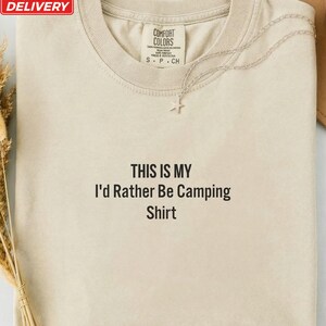 Peut inclure: Un t-shirt à col rond couleur ivoire avec le texte imprimé en noir 'THIS IS MY I'd Rather Be Camping Shirt' centré sur la poitrine. Le t-shirt présente une étiquette de marque Comfort Colors à l'encolure et un collier étoile argenté.