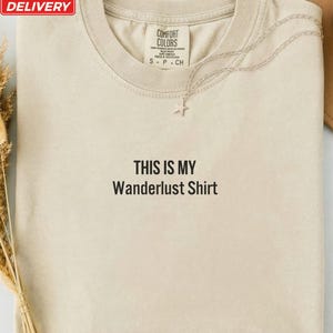 May include: An ivory-colored crew neck t-shirt featuring the black embroidered text "THIS IS MY Wanderlust Shirt" centered on the chest. The garment includes a Comfort Colors brand label at the inner neckline and a delicate silver star pendant necklace.