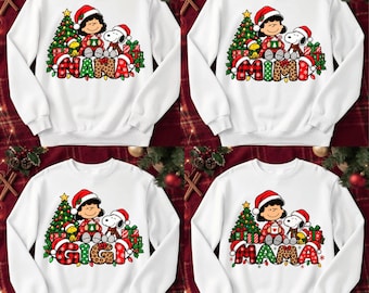 May include: Four white crewneck sweatshirts featuring a graphic of Lucy and Snoopy from Peanuts sitting by a Christmas tree with presents. Each shirt displays a different name in large, patterned letters: NANA, MIMI, GIGI, and MAMA.