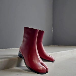 Puede incluir: Un par de botines de cuero rojo con diseño de puntera dividida. Los botines tienen un tacón bajo negro y un acabado liso y pulido. Están sobre una superficie gris clara.
