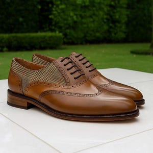 Puede incluir: Un par de zapatos de vestir de cuero marrón con diseño de ala. Los zapatos tienen un panel de tela a cuadros y cordones marrones oscuros. Se exhiben sobre una superficie blanca con un fondo verde.