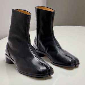 Puede incluir: Un par de botines de cuero negro con diseño de punta dividida. Los botines tienen un tacón bajo y un acabado liso y pulido. El forro interior es de color beige claro. Los botines se muestran sobre una superficie blanca.