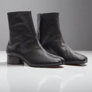 Puede incluir: Un par de botines de cuero negro con un diseño único de punta dividida. Los botines tienen un tacón de bloque de altura media y un acabado liso. Los botines miden aproximadamente 18 cm de altura.