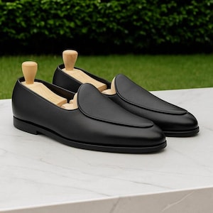Puede incluir: Un par de mocasines de cuero negro con hormas de madera. Los zapatos tienen una punta redondeada y un tacón bajo. Los mocasines se exhiben sobre una superficie de mármol blanco con un fondo verde.