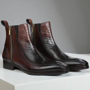 Puede incluir: Botines de cuero marrón con un patrón texturizado, similar a la piel de reptil, en la parte superior. Las botas tienen una cremallera lateral con un tirador dorado y un tacón bajo. La puntera de la bota tiene un patrón de costura decorativo.