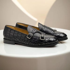 Puede incluir: Un par de zapatos de vestir de cuero negro con un patrón texturizado similar al de un cocodrilo. Los zapatos presentan correas de doble hebilla con hebillas plateadas y un diseño elegante. Los zapatos se exhiben sobre una superficie blanca.