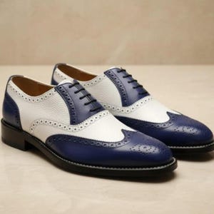 Puede incluir: Un par de zapatos de vestir de cuero bicolor en blanco y azul marino. Los zapatos presentan un diseño clásico de ala con perforaciones decorativas. Cordones negros. Ideales para un look elegante.