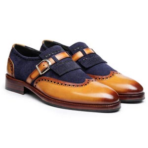 Puede incluir: Un par de zapatos de cuero de dos tonos. Los zapatos presentan una pala de cuero marrón claro y una parte superior de ante azul marino con una correa decorativa y una hebilla. Los zapatos tienen suela y tacón marrones.