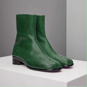 Puede incluir: Botines de cuero verde con diseño de punta dividida. Los botines tienen un tacón bajo negro y una silueta elegante y moderna. Los botines se exhiben sobre una superficie blanca.
