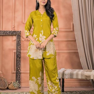 May include: A woman in a two-piece olive green pajama set. The set features large white floral patterns. The top is a button-down shirt with three-quarter sleeves. The trousers are wide-legged.