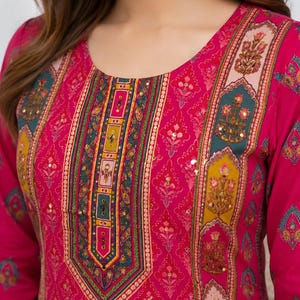 May include: A bright pink long-sleeved tunic featuring a round neckline and a central vertical panel with intricate floral and geometric patterns in teal, yellow, and gold. The fabric is adorned with small gold sequins and a repeating floral print.