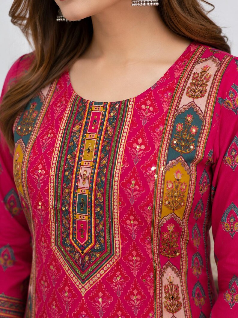 May include: A bright pink long-sleeved tunic featuring a round neckline and a central vertical panel with intricate floral and geometric patterns in teal, yellow, and gold. The fabric is adorned with small gold sequins and a repeating floral print.