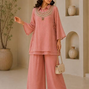 May include: A dusty rose tunic and wide-leg trousers set. The tunic features a collared neckline, flared sleeves, side drawstring ties, and a decorative embroidered yoke with metallic accents. The trousers are loose-fitting with a matching solid colour.