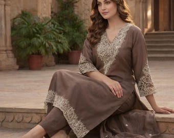 May include: A brown embroidered kurta set. The set features a long-sleeved top with white embroidery on the neckline and sleeves, matching trousers, and a sheer scarf. The outfit is worn with beige flats.