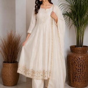May include: An off-white A-line kurta dress featuring three-quarter sleeves, a round neckline, and gold embroidery along the hem. The outfit includes matching straight-leg trousers and a sheer dupatta with a gold-trimmed scalloped edge.