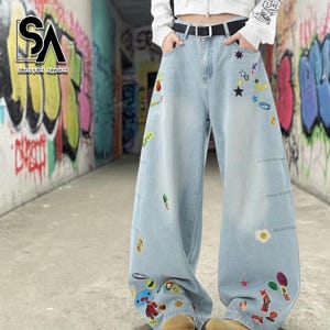 May include: Light blue denim wide-leg jeans with colorful embroidered patches. The patches include stars, food items, and cartoon characters. The jeans have a black belt and pockets. The brand logo "SA Sherrydel Apparel" is visible in the upper left corner.