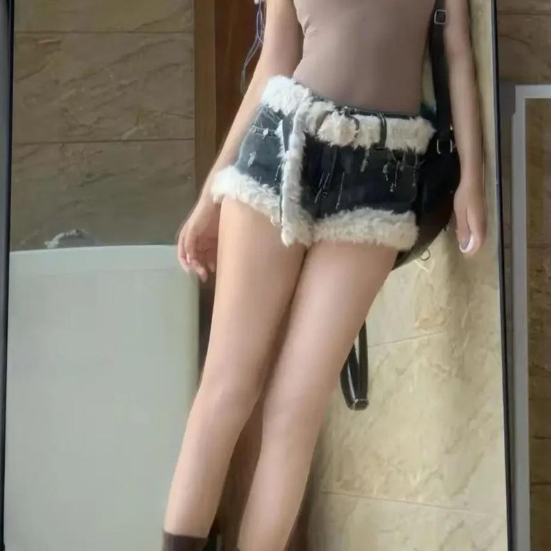 May include: Dark wash denim shorts with a cream-colored faux fur trim. The shorts have a high waist and a belt. The model is wearing a brown top and brown boots. The shorts are a trendy fashion item.