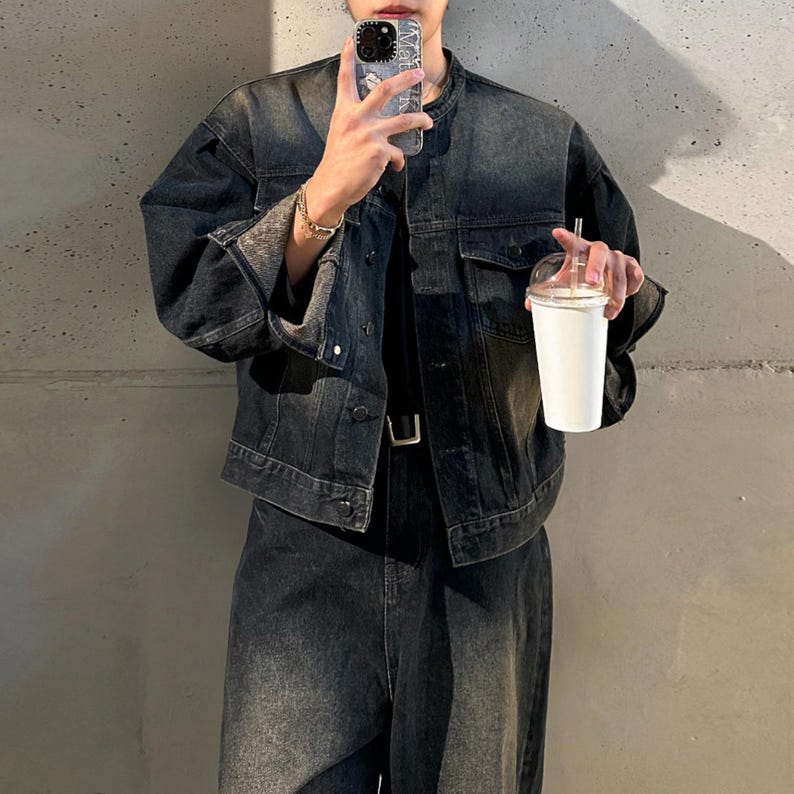 May include: A dark-wash denim jacket with a faded, worn appearance. The jacket features a classic collar, button-down front, and two chest pockets. The model is wearing a matching pair of jeans and holding a white cup with a clear lid.