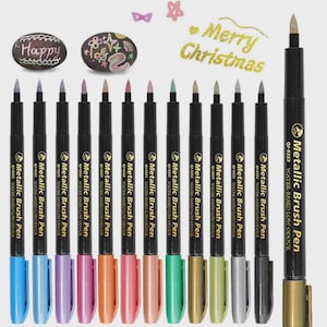 Pode incluir: Um conjunto de canetas de pincel metálicas em várias cores, com a inscrição "Metallic Brush Pen" e "Water-Based Low Odour". A imagem também inclui duas pedras decoradas e a frase "Merry Christmas".