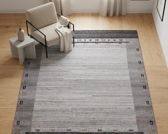 May include: A rectangular area rug featuring a light gray center panel framed by a darker gray border. The border includes horizontal black lines and small geometric symbols in black and muted brown tones. The rug has a textured, woven appearance.