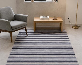 Puede incluir: Una alfombra rectangular con un patrón de rayas horizontales en varios tonos de gris carbón, gris claro y blanco. La alfombra tiene una apariencia texturizada con un detalle de flecos cortos a lo largo de los bordes más cortos, cubriendo el suelo.