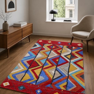 May include: A rectangular area rug featuring a vibrant geometric pattern with bold zig-zag lines in red, blue, yellow, and beige. The design includes a solid red border at the top and bottom edges, accented with small, colorful diamond-shaped motifs.