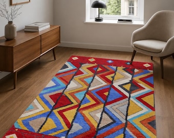 May include: A rectangular area rug featuring a vibrant geometric pattern with bold zig-zag lines in red, blue, yellow, and beige. The design includes a solid red border at the top and bottom edges, accented with small, colorful diamond-shaped motifs.