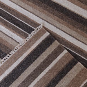 Puede incluir: Una alfombra tejida con un patr&oacute;n repetitivo de rayas verticales en tonos marr&oacute;n oscuro, marr&oacute;n claro, beige y blanco roto. La alfombra tiene un borde visible con un detalle de flecos cortos y anudados en el lateral, mostrando un tejido texturizado.