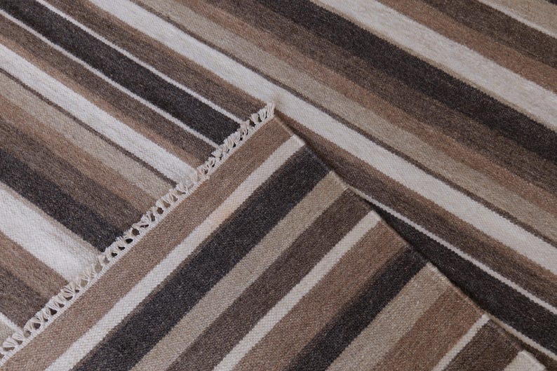 Puede incluir: Una alfombra tejida con un patr&oacute;n repetitivo de rayas verticales en tonos marr&oacute;n oscuro, marr&oacute;n claro, beige y blanco roto. La alfombra tiene un borde visible con un detalle de flecos cortos y anudados en el lateral, mostrando un tejido texturizado.