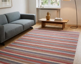 Puede incluir: Una alfombra rectangular con un patrón de rayas horizontales repetitivas en tonos de rojo apagado, azul, bronceado y crema. La alfombra tiene una textura tejida plana y está terminada con detalles de flecos blancos cortos en los dos bordes cortos.