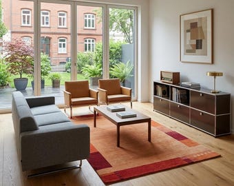 May include: A living room featuring a gray sofa, two tan leather armchairs, and a wooden coffee table on an orange and red patterned rug. A brown modular storage unit holds a radio and books, with a gold lamp and framed abstract art nearby.