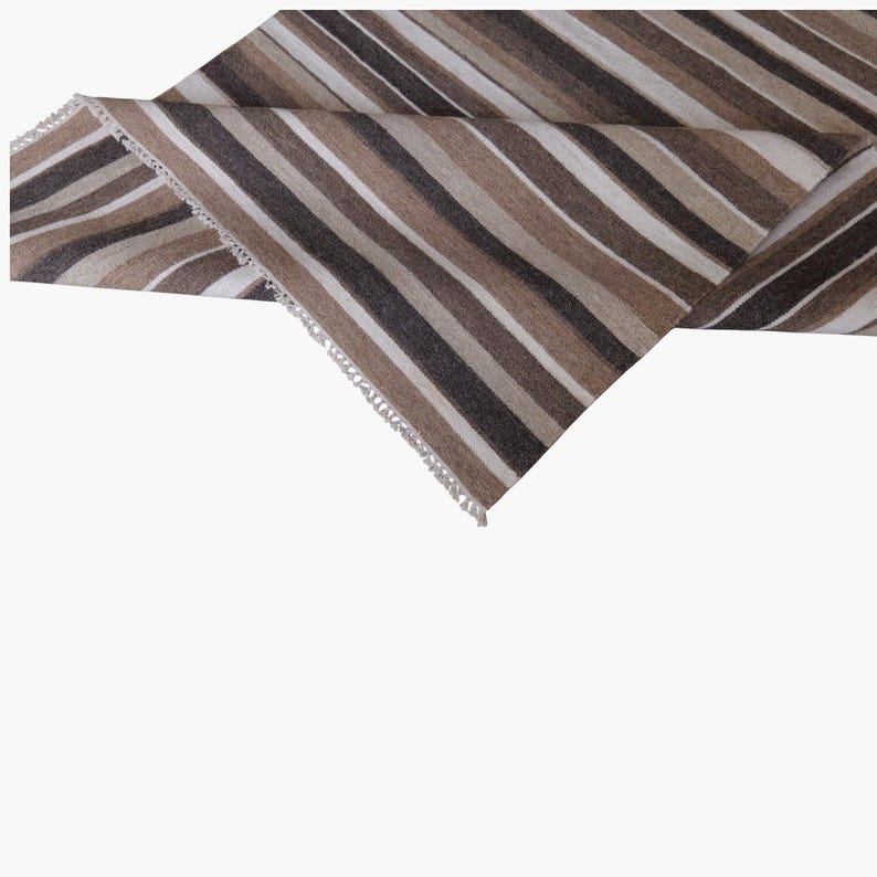 Puede incluir: Un camino de mesa rectangular de tela con un patr&oacute;n repetitivo de rayas verticales en tonos marr&oacute;n oscuro, marr&oacute;n claro, beige y blanco. La tela tiene una textura tejida visible y est&aacute; rematada con un delicado fleco blanco en los bordes.