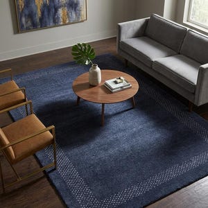 May include: A large rectangular area rug in deep navy blue featuring a white dotted border pattern. The rug is positioned under a round wooden coffee table, two tan leather chairs with gold frames, and a gray sofa on a dark wood floor.