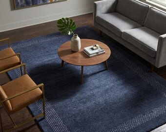 May include: A large rectangular area rug in deep navy blue featuring a white dotted border pattern. The rug is positioned under a round wooden coffee table, two tan leather chairs with gold frames, and a gray sofa on a dark wood floor.