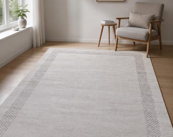 May include: A rectangular area rug featuring a light beige center and a darker gray border with a repeating dotted pattern. The rug has a flat-woven texture and covers a portion of the wooden floor, extending from the foreground toward the back.