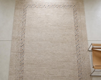 May include: A rectangular area rug in a neutral beige tone featuring a decorative border composed of small, multi-colored square patterns in shades of blue, brown, and grey, set against a solid, textured, light-colored central field.