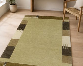 May include: A rectangular area rug featuring a large solid sage green center panel surrounded by a border of alternating blocks in shades of dark brown, olive green, tan, and cream, accented with small linear stitch-like patterns.