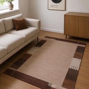 Peut inclure: Un tapis rectangulaire doté d'un grand panneau central beige uni, encadré par une large bordure composée de blocs alternés marron foncé, marron moyen et beige clair, agrémenté de motifs de lignes parallèles dans les coins.