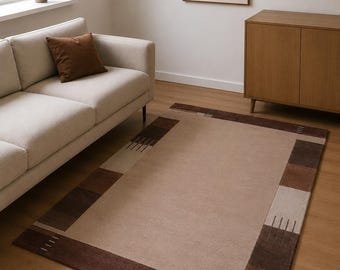 May include: A rectangular area rug featuring a large solid beige center panel framed by a wide border of alternating dark brown, medium brown, and light beige blocks, accented with thin, parallel line patterns in the corner sections.