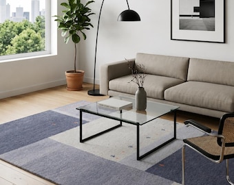 May include: A rectangular area rug featuring a geometric block pattern in shades of blue, gray, and cream. The rug has a textured surface with small, scattered embroidered details in orange and blue, covering a large portion of the wooden floor.