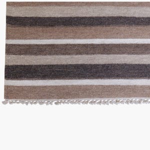 Puede incluir: Una alfombra tejida con rayas horizontales en varios tonos de marr&oacute;n, beige y blanco. El tejido tiene una apariencia texturizada con un fleco corto y anudado a lo largo del borde inferior, creando un patr&oacute;n consistente en la superficie.