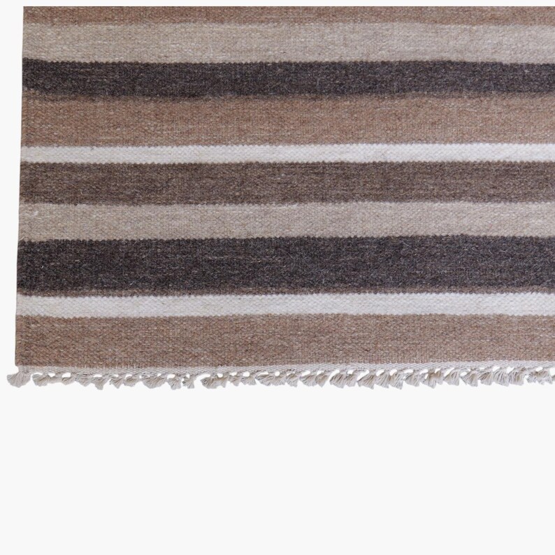 Puede incluir: Una alfombra tejida con rayas horizontales en varios tonos de marr&oacute;n, beige y blanco. El tejido tiene una apariencia texturizada con un fleco corto y anudado a lo largo del borde inferior, creando un patr&oacute;n consistente en la superficie.