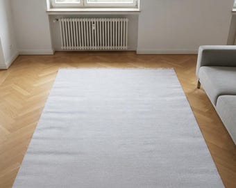May include: A rectangular light gray area rug with a flat-woven texture and short fringe detailing along the top and bottom edges. The rug is positioned centrally on a light brown wooden floor with a herringbone pattern, featuring a solid color.
